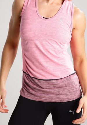 Kobieta ubrana w różowy, sportowy top bez rękawów i czarne legginsy marki Puma, stojąca z lekko ugiętymi ramionami na jednolitym tle.