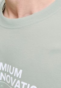 Šviesiai žalia medvilninė marškinėliai su apykakle, su balta spausdinta užrašu "PREMIUM INNOVATION" ant priekio, lygi tekstūra.