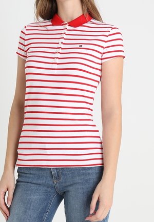 Woman wearing a fitted white polo shirt with red horizontal stripes and a red collar, paired with blue denim jeans.