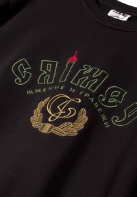 Zwarte sweatshirt met groene en rode geborduurde tekst. Heeft een gouden lauwerkrans ontwerp en een emblematische illustratie bij de halslijn.