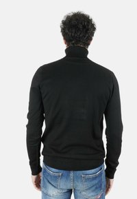 Uomo con capelli ricci che indossa un maglione a collo alto nero e jeans blu, visto di spalle su uno sfondo bianco uniforme.