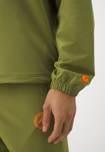 Olive green athletic top with elastic cuff. Features a round orange button and a circular orange logo on the lower garment. Smooth texture.