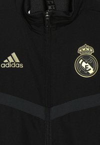 Black zipped jacket with gold Adidas logo on left and gold Real Madrid football club crest on right chest.