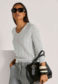 Frau mit großen schwarzen Sonnenbrillen, einem hellblauen Strickpullover, grauen Hosen, die eine schwarze Handtasche hält und dicke Armbänder am Handgelenk trägt.