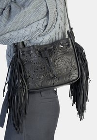 Czarna skórzana torebka z misternym wzorem z ćwieków w kształcie zawirowań, frędzlami na krawędziach i zamknięciem na sznurek. Noszona z szarym swetrem i spodniami.