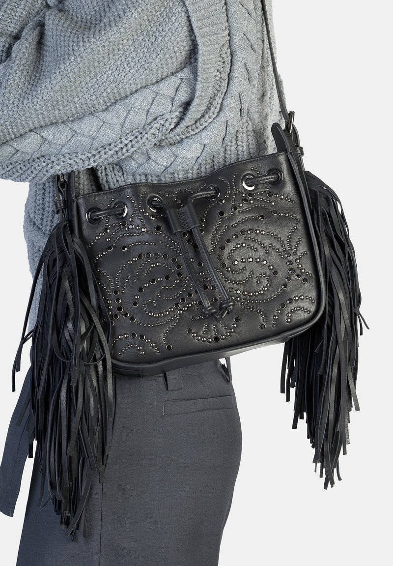 Czarna skórzana torebka z misternym wzorem z ćwieków w kształcie zawirowań, frędzlami na krawędziach i zamknięciem na sznurek. Noszona z szarym swetrem i spodniami.