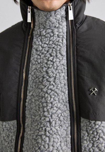 Svart ytterjacka med en grå texturerad fleeceframsida, två blixtlås och en liten metallisk hammarsymbol på vänster bröst.