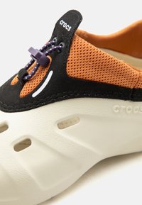 Chaussure Crocs avec tissu en mesh orange, zone lacets noire, lacets élastiques violets avec stoppeur et semelle en mousse beige ventilée.