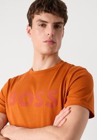 Oransje bomulls t-skjorte med rund hals, med en fremtredende rød "BOSS" logo over brystet. Glatt tekstur og standard passform.