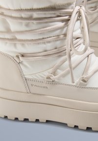 Beżowy ocieplany but z teksturowaną gumową podeszwą, posiadający pikowany design i kontrastujące sznurowadła. Lekka tkanina i branding Primaloft.