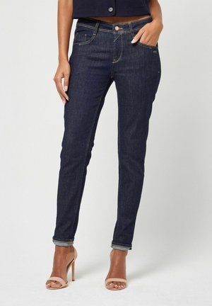 Frau trägt dunkelblaue, hochgekrempelte Skinny-Jeans und beige Sandalen mit hohen Absätzen und steht vor einem schlichten Hintergrund.