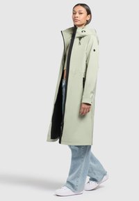 Long, light green raincoat with a hood and black zipper detailing. Worn over a cropped top and wide-leg light blue jeans with white sneakers.