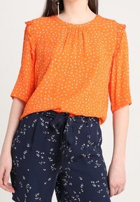 Orange bluse med hvidt bladmønster, korte flæseærmer, rund halsudskæring, parret med marineblå bukser med små blomsterprint og bindebånd i taljen.