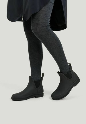 Person trägt dunkelgraue Leggings, schwarze knöchelhohe Regenstiefel und einen schwarzen Mantel und steht vor einem hellen Hintergrund.