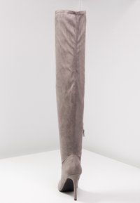 Bota alta de ante gris hasta el muslo con punta afilada y tacón alto de aguja. Cuenta con un cierre de cremallera en el lateral exterior para facilitar su uso.