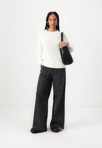 Übergroßer weißer Strickpullover und schwarze weit geschnittene Hose mit silbernem Punktmuster, kombiniert mit einer schwarzen Schultertasche und klobigen schwarzen Schuhen.
