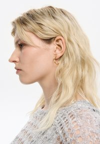 Goldene Creolen mit gedrehtem Design, die eine glänzende Textur aufweisen. Das Model hat welliges, blondes Haar und trägt ein silbernes Strickoberteil.