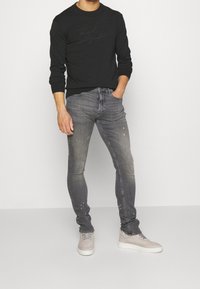 Schwarzer Sweatshirt mit einem strukturierten Logo, kombiniert mit schmal geschnittenen, ausgewaschenen grauen Jeans mit leichten Spritzdetails und hellgrauen Sneakern.