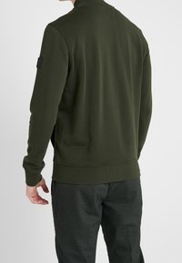 Tmavě zelená mikina s žebrovaným límcem a manžetami, s texturovanou látkou a nášivkou na levém rukávu. Nošeno se kostkovanými kalhotami.