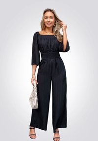 Mono negro con escote cuadrado, cintura fruncida y piernas anchas. Combinado con un bolso de recolección beige claro y sandalias negras con tiras.