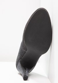 Chaussure noire à talon haut pointant vers le haut, avec une semelle texturée, un talon étroit et un dessus en tissu pailleté, placée sur des surfaces blanches.