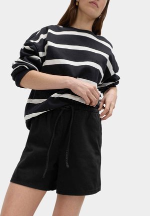Vrouw die zwarte shorts met trekkoord verstelth, draagt een zwart-wit gestreept shirt met lange mouwen en gouden oorbellen, tegen een effen achtergrond.