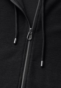 Sudadera con cremallera negra, de tela texturizada, con una cremallera plateada y cordones con puntas de metal, vista en primer plano.