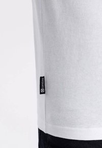 Vit bomull T-shirt med kort ärm, med en liten svart etikett som står "ombre" på sömmen. Slät textur, avslappnad design.