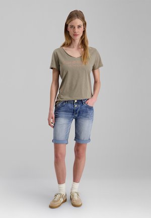 Woman wearing khaki short-sleeve shirt with text, knee-length rolled denim shorts, cream socks, and beige sneakers standing on gray background.