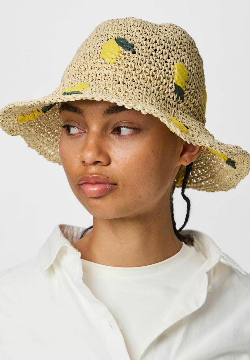 Femme portant un chapeau bob beige crocheté avec une broderie florale jaune, une chemise blanche, regardant vers sa gauche sur un fond uni.