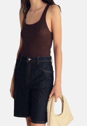 Kvinde iført en brun, ærmeløs ribbet top og mørke denimshorts, der holder en tekstureret beige vævet håndtaske i højre hånd.