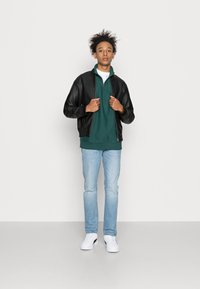 Casaco bomber de couro preto sobre um sweatshirt verde com fecho, combinado com jeans azul claro e sapatilhas brancas. Conjunto casual, padrão minimalista.