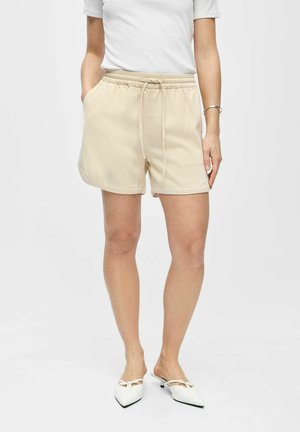 Femme portant un short beige à taille élastique avec cordon de serrage, un haut blanc à manches courtes, et des chaussures blanches à talons bas pointus avec des nœuds.