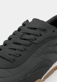 Sneaker nera con tomaia in pelle texturizzata, lacci grigi piatti e dettaglio del logo discreto. Presenta una suola in gomma marrone chiaro a contrasto e una finitura liscia.