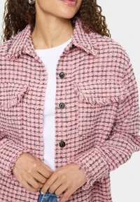 Roza tweed jakna s črno-belo šahovsko vzorcem, raztrganimi robovi in črnimi gumbi. Nosi se čez belo srajco, ki prikazuje sprednje žepke.