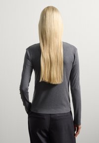 Haut gris à manches longues avec une coupe ajustée, fabriqué en tissu doux. Porté avec un pantalon sombre, vu de dos, avec des cheveux blonds raides.