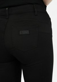 Fekete elasztikus farmernadrág, slim fit szabással, hátsó zsebbel, amelyen fekete címke található, valamint finom varrási részletekkel.