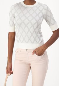 Mujer vistiendo un suéter corto de manga blanca con un patrón de diamantes y pantalones de un rosa pálido, con una mano en el bolsillo y sosteniendo una correa beige con la otra mano.