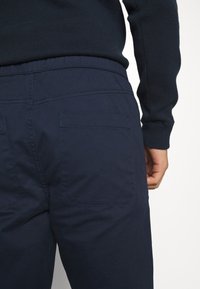 Mörkblå byxor med elastisk midja, har två bakfickor och subtila sömdetaljer. Slät, lätt tygtextur.