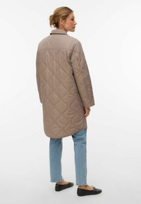 Naine heledast pruunist tikandiga pika jope, heleroheliste teksade ja mustade baleriinide sees, seisab veidi vasakule suunaga valgel taustal.