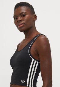 Svart atletisk crop-top med hvit tre-stripes mønster langs sidene. Har rund hals og et tettsittende design.