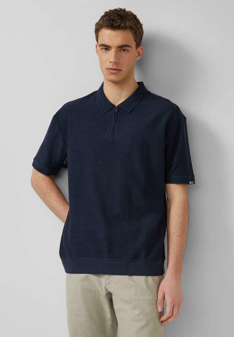 Junger Mann mit kurzen lockigen Haaren, der ein marineblaues Poloshirt mit kurzen Ärmeln und hellbeige Hosen trägt, steht vor einer grauen Wand.