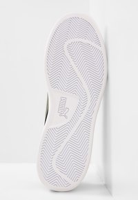 Suela de zapatilla Puma con una base de goma blanca, patrón de banda de rodadura en zigzag y detalle del logo. La parte superior del zapato es predominantemente verde.
