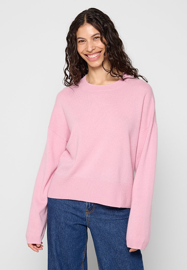 Różowy, długi sweter z długim rękawem o okrągłym dekolcie i luznym kroju. Miękka, teksturowana tkanina w zestawieniu z ciemnoniebieskimi spodniami o szerokich nogawkach.