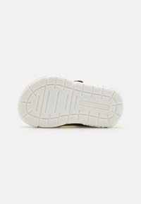 Svart sneakers med en hvit gummisåle som har et teksturert grep mønster; inkluderer gule detaljer og logo på siden.