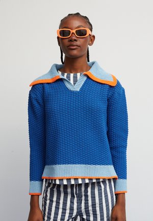 Μπλε πλεκτή μπλούζα με ανάγλυφη υφή, λευκό γαλάζιο γιακά και πορτοκαλί λεπτομέρειες. Συνδυασμένη με ριγέ, πλισέ παντελόνια και πορτοκαλί γυαλιά ηλίου.
