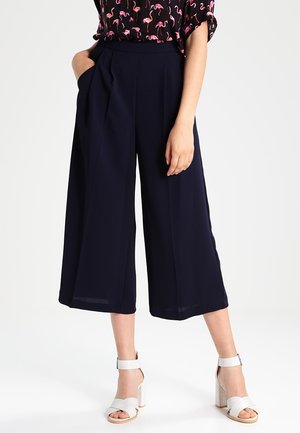 Kobieta w granatowych szerokich spodniach o skróconej długości, czarnym topie z różowym nadrukiem flaminga i białych sandałach na wysokim obcasie, stojąca z jedną ręką w kieszeni.