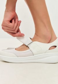 White shoe featuring a breathable mesh upper and a velcro strap. The design includes a wedge sole with rounded edges and cut-out accents.