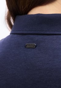 Κοντινό πλάνο σε γιακά πουκάμισου από υφασμάτινη ύλη σε σκούρο μπλε με υφή, με μια μικρή μεταλλική ετικέτα ραμμένη στο πίσω μέρος, που εμφανίζει το όνομα της μάρκας "Barbour".