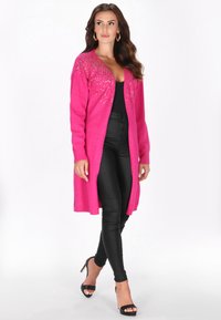 Egy nő élénk rózsaszín hosszú kardigánt visel díszítésekkel, fekete felsőt, fekete testhez simuló nadrágot és fekete magas sarkú szandált, magabiztosan sétál.